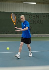 Tennis spiller iført en blå polo-shirt og sorte shorts, som holder en orange ketsjer, klar til at slå en gul bold på en blå bane.