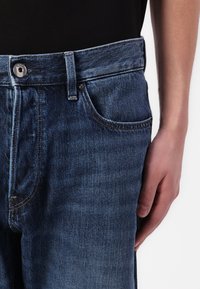 Jeans de mezclilla azul oscuro con corte recto, que cuentan con cierre de botón, pasadores para cinturón y bolsillos frontales. La tela tiene una ligera textura.