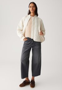 Veste bomber matelassée beige clair, pull beige en peluche, jeans large gris foncé et baskets en daim marron. Composition de tenue simple et décontractée.