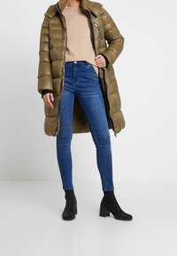 Olivgrüner gefütterter Mantel mit Reißverschluss und Kapuze, kombiniert mit blauen Skinny Jeans mit Steppdetails und schwarzen, gepolsterten Ankle Boots.