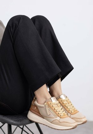 Zapatillas beige y marrón con un acabado texturizado, con cordones en la parte delantera, detalles dorados y un cierre con cremallera. Llevadas con pantalones negros ligeramente acampanados.