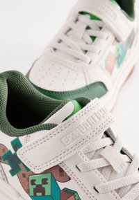 Baskets en cuir blanc avec des accents verts, doublure en mesh texturé, design à lacets, et un motif pixélisé avec des personnages en blocs.