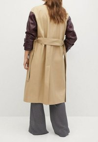 Trench beige con maniche in pelle marrone, con cintura in vita. Presenta un design di lunghezza media e una texture liscia. Pantaloni a motivi visibili sotto.