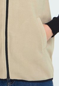 Beige Fleece-Weste mit schwarzem Reißverschluss und gerippten Bündchen, ausgestattet mit Seitentaschen und einer glatten Textur.