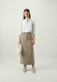 Bluză albă cu decolteu tip chei și mâneci bufante, asortată cu o fustă midi din denim bej. Modelul ține o geantă albă, purtând bocanci gri până la gleznă.