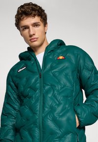 Tyrkysová péřová bunda se zapínáním na zip, která má kapuci, strukturovaný vzor a vyšívané logo v oranžové a žluté barvě na hrudi.
