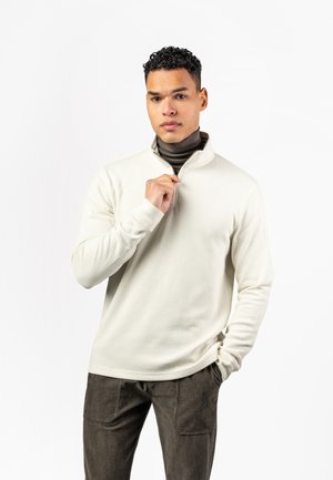 Young man with curly hair wearing a cream zip-up sweater over a dark turtleneck, standing with one hand in pocket and the other adjusting the zipper.