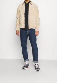 Chaqueta de pana beige con cuello tipo solapa, cierre de cremallera y dos bolsillos laterales. Se usa sobre una camisa negra de botones, con jeans azules y zapatillas.