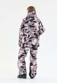 Sukelluspuvut piiloutumakuosilla harmaan ja violetin sävyissä, jossa on huppu, ruudukkomallisia yksityiskohtia ja löysä malli. Mustat kengät näkyvissä.