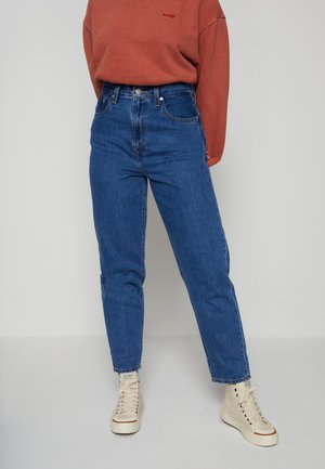 Personne portant un jean bleu taille haute, un sweat-shirt couleur rouille et des baskets montantes blanches, debout devant un fond clair uni.