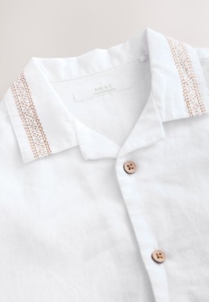 Valge linane särk pruuni nööpide ja pruuni tikandiga krae servadel, millel silt "NEXT, Made in Bangladesh."