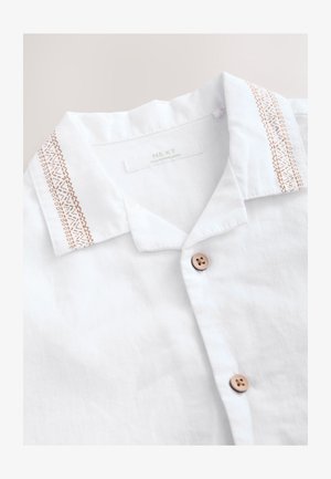 Fehér lenvászon ing barna gombokkal és barna hímzett mintákkal a gallér szélén, "NEXT, Made in Bangladesh" címkével.