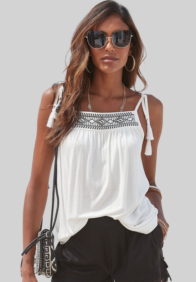 Top sin mangas blanco con diseño bordado negro en el escote, con detalles de borlas y un corte relajado, combinado con pantalones cortos negros.