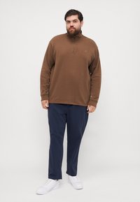 Bruine pullover met rits in een zachte stof, gecombineerd met marineblauwe broek. Model draagt witte sneakers en staat tegen een neutrale achtergrond.