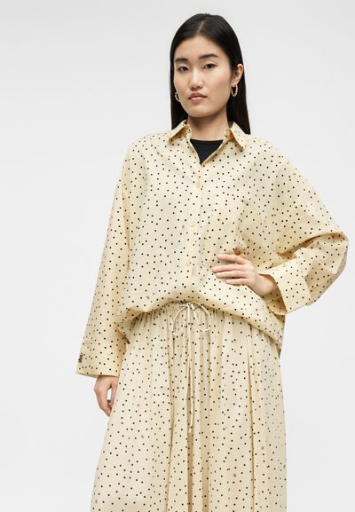 Femme aux longs cheveux bruns portant un chemisier beige à pois et une jupe assortie, posant avec une main sur la hanche sur un fond uni.