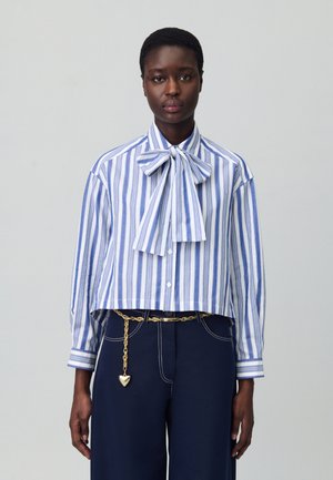 Camicia a maniche lunghe, tagliata, a righe blu e bianche, con un grande papillon al colletto. Abbinata a pantaloni blu navy e una cintura in catena dorata.