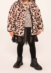 Rosa Leopardenmuster-Faux-Fur-Jacke, schwarzes und weißes gepunktetes Oberteil, schwarzer Tüllrock, schwarze Leggings und schwarze Ankle-Boots mit strukturierten Sohlen.