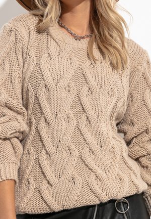 Beige neulepaita, jossa on tekstuuri kaapelikuviota, leveät hihat ja ribbireuna. Ketju koru näkyvissä kaula-aukossa.