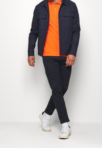 Granatowa kurtka zapinana na guziki z dwiema kieszeniami na piersi, pomarańczowa koszulka polo, granatowe spodnie oraz białe trampki z czarnym akcentem na pięcie.