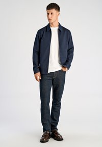 Granatowa kurtka na zamek błyskawiczny na białej koszulce, zestawiona z ciemnymi dżinsami i brązowymi eleganckimi butami. Materiał wydaje się gładki, z minimalnymi detalami designu.