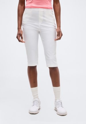 Osoba ubrana w białe spodnie do kolan, białe skarpetki, białe buty i różową koszulę na jasnym, jednolitym tle.