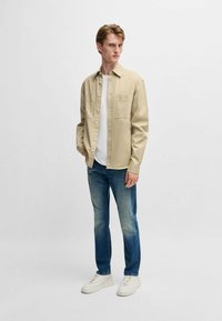 Vaalean beige nappikauluspaita, jossa on kaksi rintataskua, käytetty valkoisen t-paidan päällä, yhdistettyinä haalistuneisiin sinisiin farkkuihin ja valkoisiin tennareihin. Yksinkertainen muotoilu ja rento istuvuus.