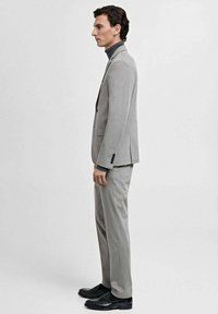 Completo grigio su misura con un design strutturato, vestibilità slim, tasche laterali e scarpe nere. Il modello indossa un dolcevita scuro sotto.