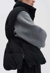 Schwarze Steppweste mit hohem Kragen und elastischem Bund. Sie verfügt über eine glatte Textur und einen seitlichen Reißverschluss, die über einem grauen Langarmkleidungsstück getragen wird.