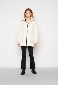 Witte pufferjack met een hoge kraag, voorkant met rits en twee grote zakken, gedragen over zwarte flared jeans en zwarte laarzen.