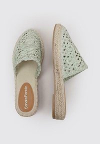 Zapatos deslizantes de tejido verde claro con la parte trasera abierta, que presentan una gruesa suela de yute trenzado y una plantilla interior lisa con el logotipo.