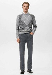 Suéter gris de rombos con diamantes blancos y grises oscuros, combinado con jeans grises y zapatos negros. El modelo está de pie contra un fondo blanco.