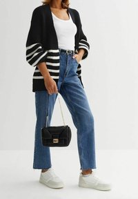 Kvinna iklädd en svart och vit randig kofta, vit linne, blå jeans, vita sneakers, hållande en liten svart handväska med en guldkedja.