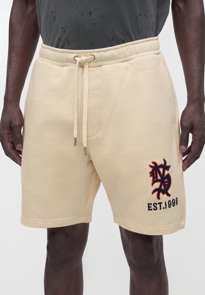Homme portant un short beige à cordon avec un logo brodé noir et rouge et le texte "EST. 1999" sur la jambe gauche, assorti à une chemise gris foncé délavée.