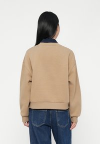 Beige genser med senket skuldre og ribbestrikkede mansjetter. Har en marineblå krage og kortdelt nederkant, brukt sammen med blå denimjeans.