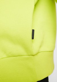 Sweatshirt giallo neon con orlo e polsini a coste, texture morbida e un piccolo tassello del marchio nero vicino alla cucitura.