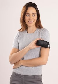 Czarna skórzana prostokątna saszetka z zamkiem błyskawicznym, o strukturalnej powierzchni i subtelnym detalem logo, trzymana przez osobę w szarym swetrze.