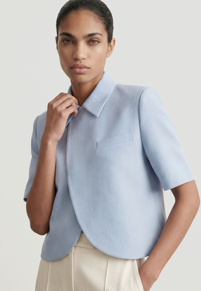 Woman with pulled-back hair wears a light blue short-sleeve jacket and cream pants, standing against a plain light background.