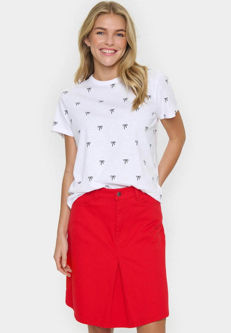 Baltā T-kreklā ar melniem loku rakstiem, apvienota ar sarkanu A-linijas svārku līdz ceļiem, kuram ir priekšējais šķēlums un pogu aizdare.