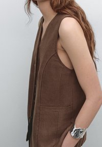 Brun ärmlös linneväst med strukturerad yta, försedd med knappar framtill och sidofickor. En silverarmbandskuff pryder armen.