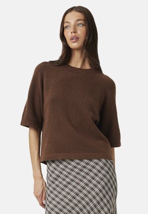 Vrouw met lang bruin haar draagt een losse bruine korte mouw trui en een zwart-witte ruiten rok, staand tegen een effen achtergrond.