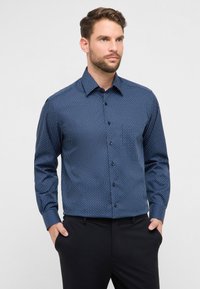 Camicia blu navy con bottoni, con un piccolo motivo geometrico bianco, maniche lunghe e colletto classico, abbinata a pantaloni neri.