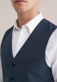 Dunkelblaues Weste aus glattem Stoff, mit Reverskragen, zwei Knöpfen und einer Brusttasche, über einem weißen, strukturierten Hemd getragen.