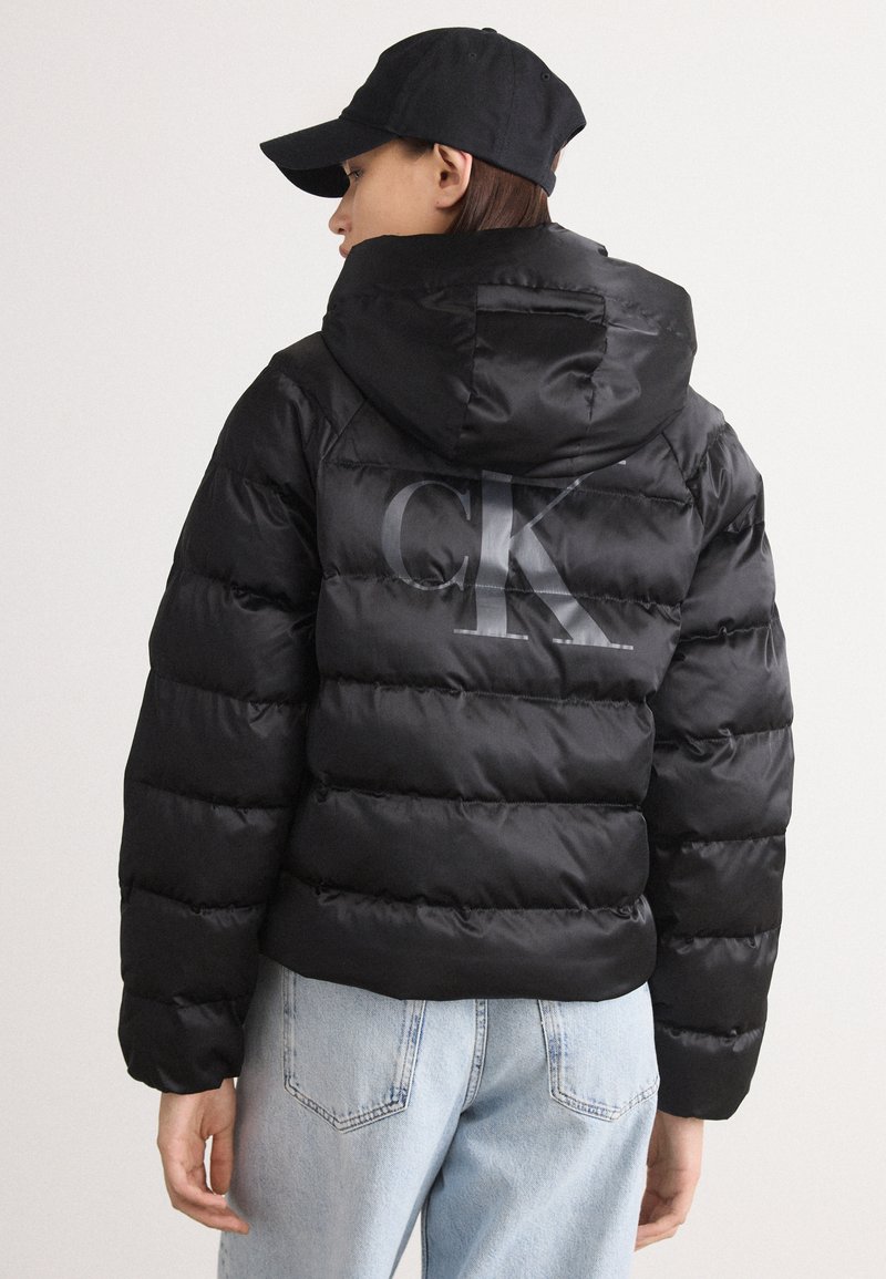 Czarna kurtka puchowa z kapturem, z błyszczącym logo "CK" na plecach. Materiał wydaje się błyszczący i teksturowany, w połączeniu z jasnoniebieskimi dżinsami.