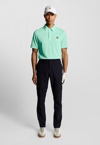 Lichtgroen polo shirt met een gele logo, marineblauwe broek, witte handschoen aan de linkerhand, witte pet en beige schoenen met een bruine zool.