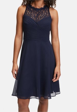 Tmavě modré šaty bez rukávů s krajkovým sedlem, plisovaným živůtkem a sukní ve tvaru A z šifonu, nošené stojící osobou.