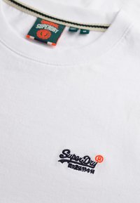 T-shirt bianco in cotone con scollatura rotonda, caratterizzato da un logo ricamato blu navy e dettagli arancioni. L'etichetta mostra il nome del marchio e i colori.