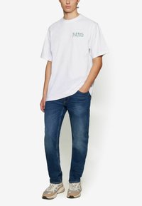 Witte katoenen T-shirt met groene bedrukte tekst, gecombineerd met blauwe denim jeans en lichtgekleurde sneakers. Casual pasvorm en recht gesneden ontwerp.