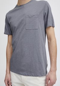 Tricou scurt cu mânecă gri, dintr-un material moale, având un buzunar pe piept, guler rotund și o croială lejeră, cu detalii minime de cusătură.