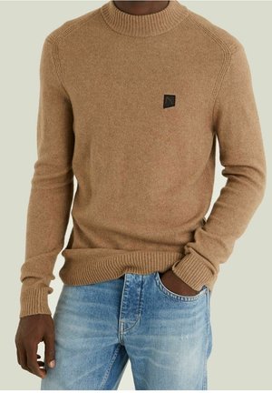 Suéter de punto marrón con un cuello y puños acanalados, que presenta un pequeño parche de logo negro en el pecho. Combinado con jeans de color azul claro.