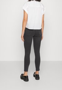 Zwarte slim-fit jeans met vijf zakken, in combinatie met een witte blouse met korte mouwen. Model staat tegen een neutrale achtergrond.
